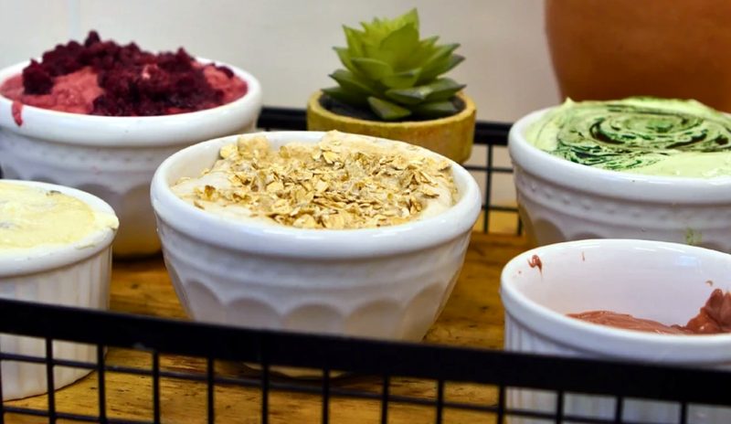 Potes brancos com pastas coloridas, uma delas com aveia por cima, sobre uma bandeja de arame preta, ao lado de uma suculenta e de uma moringa de barro