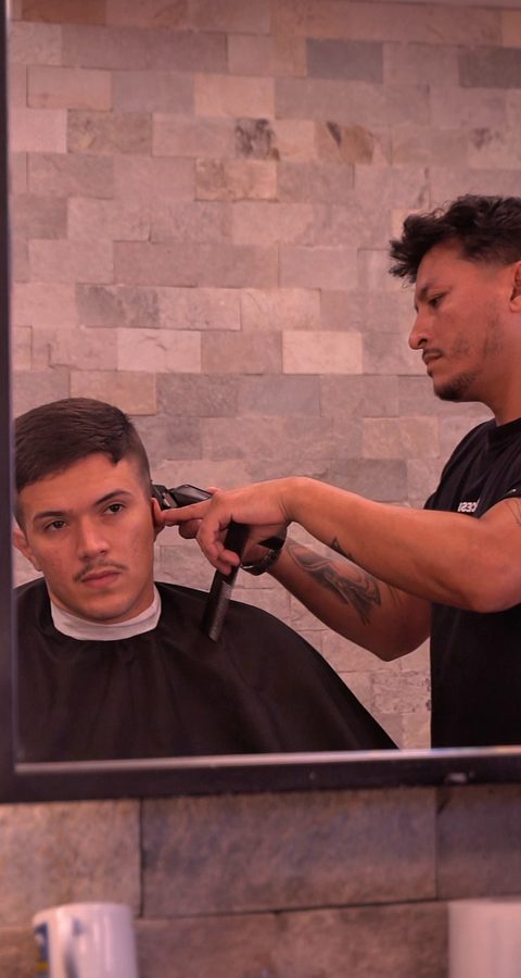 Barbeiro cortando o cabelo de um cliente diante de um espelho grande, com a parede de pedras ao fundo, na Barbearia Constant