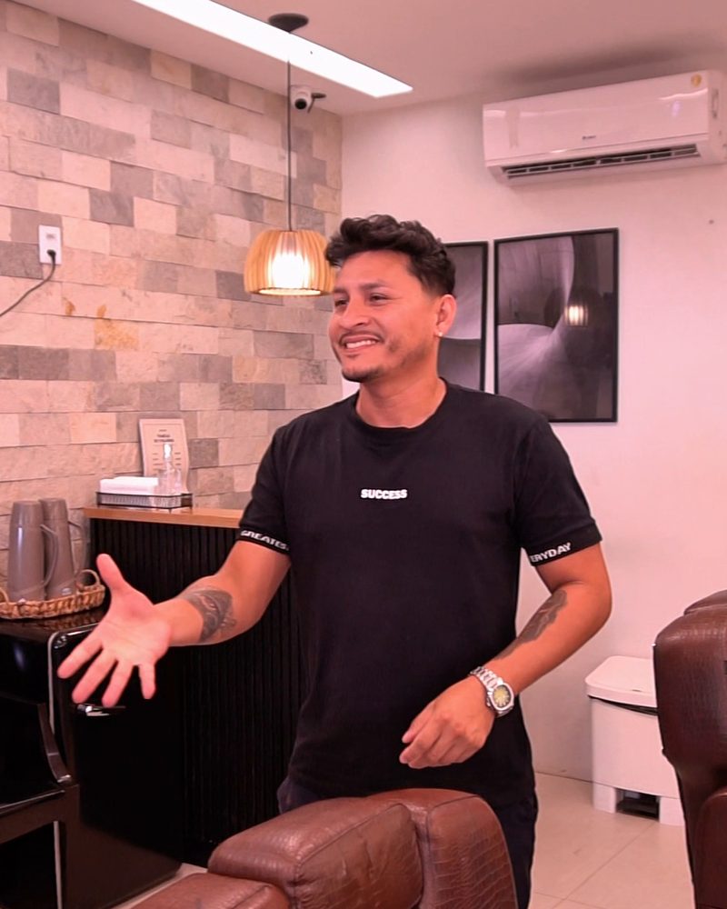 Salão da Barbearia Constant com parede de pedras, balcão preto ripado, luminária de palha e poltronas de couro marrom, com um barbeiro recebendo os clientes de braços abertos