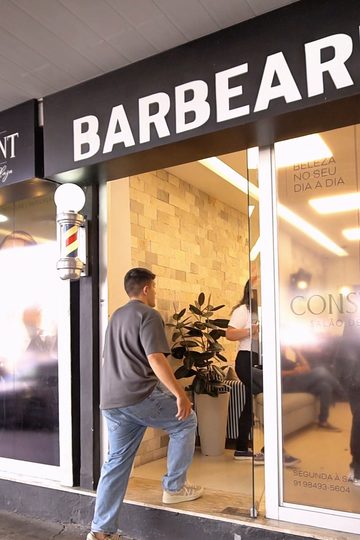 Fachada da Barbearia Constant, com o letreiro preto, a vitrine de vidro e a parede de pedras visível lá dentro
