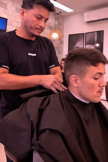 Barbeiro colocando a capa em um cliente sentado na poltrona, sob a luminária de palha, na Barbearia Constant