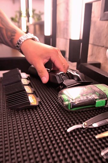 Máquinas de corte, pentes-guia e navalha sobre o tapete preto da bancada, com o espelho de luz ao fundo, na Barbearia Constant
