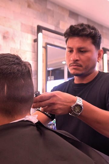 Barbeiro concentrado cortando o cabelo de um cliente, com espelhos de luz e a parede de pedras ao fundo, na Barbearia Constant