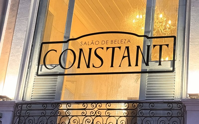 Janela em arco do casarão do Constant Benjamin à noite, acesa em tom dourado, com o nome Salão de Beleza Constant no vidro, o lustre ao fundo e a grade de ferro trabalhado da sacada