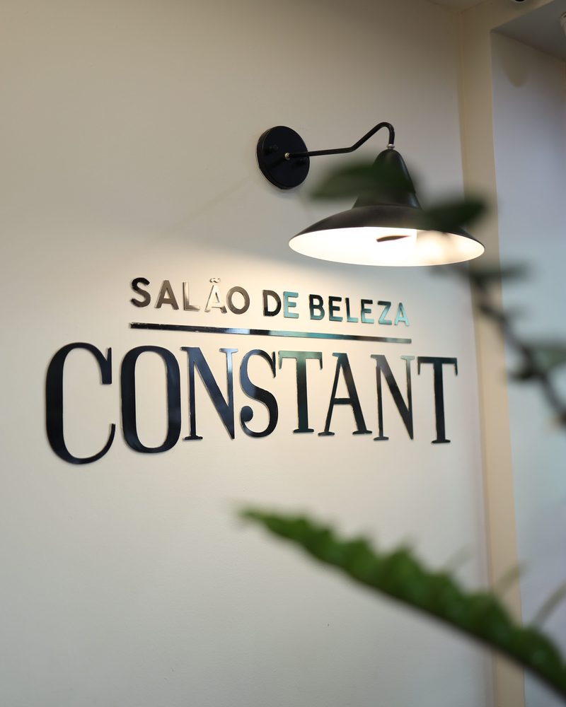 Letreiro preto com o nome Salão de Beleza Constant na parede branca do Constant Plaza, sob uma luminária de braço